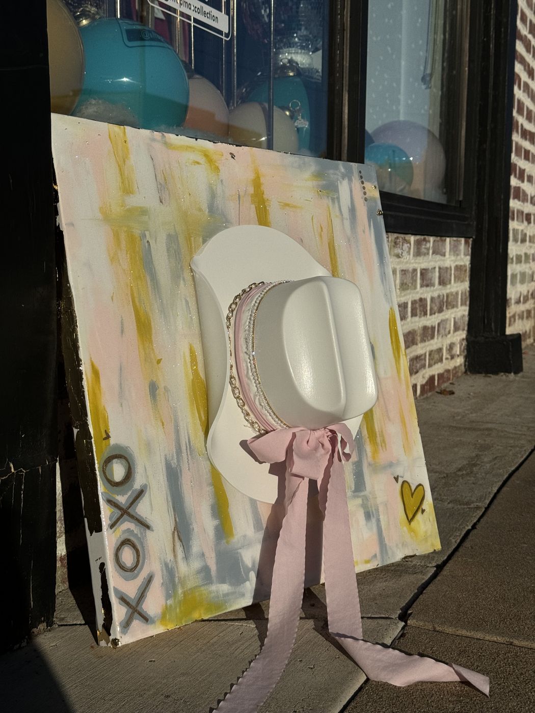 XOXO canvas with a white cowboy hat and pink ribbon, propped outside a shop window