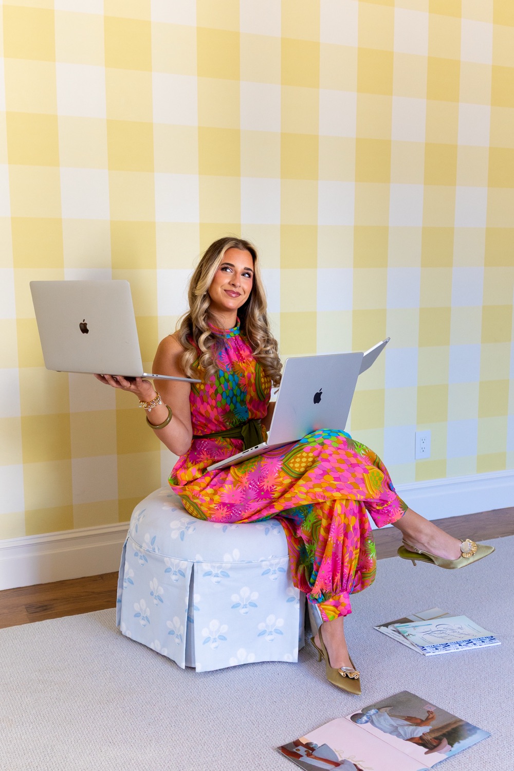 Lenzie Berry sitting on a skirted ottoman in front of yellow gingham wallpaper, holding two laptops